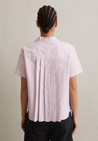 Korte mouw shirt in lichtroze met witte strepen, met een geplooid ontwerp aan de achterkant en een kraag. Stof lijkt glad en lichtgewicht te zijn.