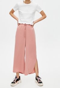 Chemise blanche courte et pantalon rose large avec fentes latérales, associés à des baskets noir et blanc avec des accents colorés.