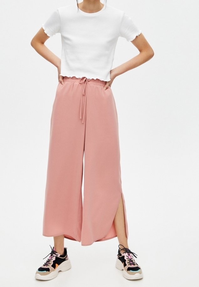 Chemise blanche courte et pantalon rose large avec fentes latérales, associés à des baskets noir et blanc avec des accents colorés.