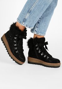 Botas negras hasta el tobillo con ribetes de pelo sintético, cordones decorativos y acentos de strass, combinadas con jeans azul claro con remangue.