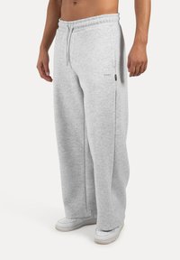 Pantalones de chándal sueltos en gris claro, con cinturilla elástica, cordón, bolsillos laterales y un pequeño detalle de logo en la cadera.