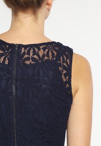 Navy blue lace dress with floral patterns, featuring a round neckline and a zip closure at the back. Textured fabric with sheer elements.