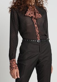 Durchsichtige schwarze Bluse mit rosa Polka-Dots und Akzenten im Leopardenmuster, mit einem Schleifenkragen und kombiniert mit gestreiften Hosen und einem Gürtel.