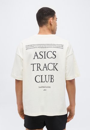 Férfi, aki fehér, túlméretezett pólót visel nagy fekete "ASICS TRACK CLUB" felirattal és kisebb motivációs szöveggel a hátán, fekete rövidnadrággal párosítva.