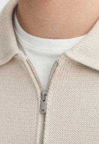 Beżowy dzianinowy sweter zapinany na zamek błyskawiczny z kontrastującą białą warstwą wewnętrzną. Posiada metalowy zamek i prążkowany kołnierz, podkreślając teksturę i design.