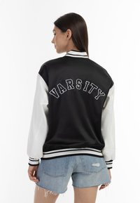 Jachetă de tip varsity, alb-negru, cu un finisaj satinat, având un imprimit mare "VARSITY" pe spate, manșete cu nervuri și un design scurt.