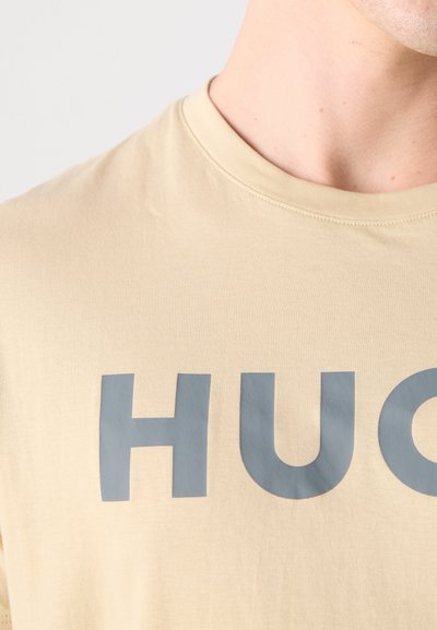 Бежева тениска от мек плат с големи сиви букви, написващи "HUG". Прост дизайн с кръгло деколте и гладка текстура, без видими шарки.