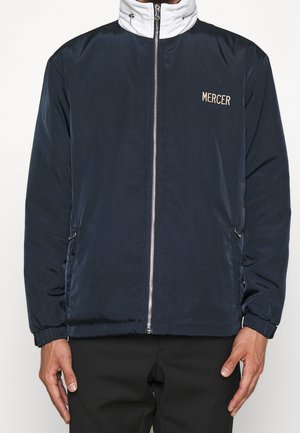 Marineblaue Reißverschlussjacke mit weißem Kragen-Detail, elastischen Bündchen, seitlichen Reißverschlusstaschen und "MERCER" auf der linken Brust gestickt.