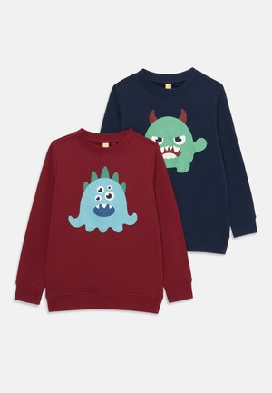Dos sudaderas: una de color burdeos con un gráfico de un monstruo azul; otra de color marino con un gráfico de un monstruo verde. Ambas tienen puños y cuello acanalados.