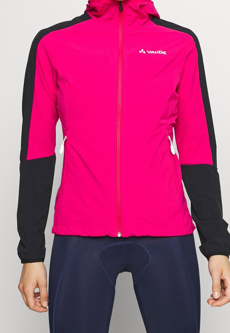 Femme portant une veste de sport zippée rose vif et noire avec des tirettes de fermeture éclair blanches et un pantalon de cyclisme bleu foncé.