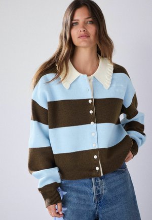 Frau mit langen braunen Haaren, die eine blau-braun gestreifte, geknöpfte Strickjacke mit einem cremefarbenen Rüschenkragen und blaue Jeans trägt, steht vor einem einfarbigen Hintergrund.