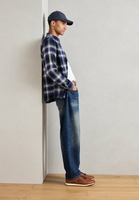 Tenue décontractée avec une chemise à carreaux bleue et jaune, un t-shirt blanc, un jean bleu délavé et des baskets marron avec des lacets foncés.