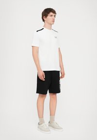 White short-sleeve t-shirt with black accents on shoulders, logo on left chest, paired with black shorts featuring a side graphic design.