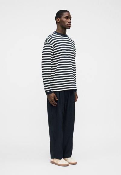 Άνδρας που στέκεται πλαγίως, φοράει μακρυμάνικο πουκάμισο με μαύρες και λευκές ρίγες, χαλαρό μαύρο παντελόνι και λευκά αθλητικά παπούτσια σε απλό φόντο.