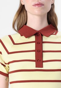 Gul polo-shirt med horisontale røde striper, en rød krage, og en knappet strop med kontrasterende knappedetaljer. Myk tekstur.