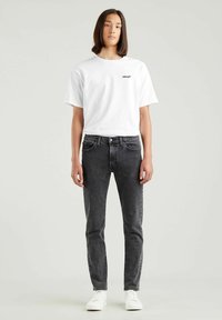 T-shirt bianca in cotone con logo nero Levi's, abbinata a jeans neri slim-fit e sneaker bianche. Design a taglio dritto, stile minimalista.