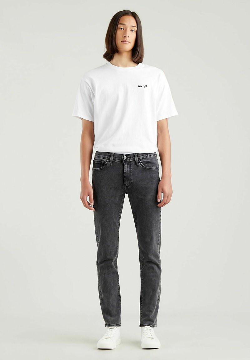 T-shirt bianca in cotone con logo nero Levi's, abbinata a jeans neri slim-fit e sneaker bianche. Design a taglio dritto, stile minimalista.