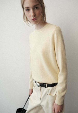 Joven mujer con un suéter amarillo claro y pantalones crema con cinturón negro, sosteniendo un bolso negro frente a un fondo de pared lisa.