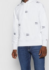 Witte hoodie met marineblauwe logopatches, voorzien van een capuchon met trekkoord, geribbelde manchetten en zoom. Gecombineerd met een blauwe spijkerbroek voor een casual uitstraling.
