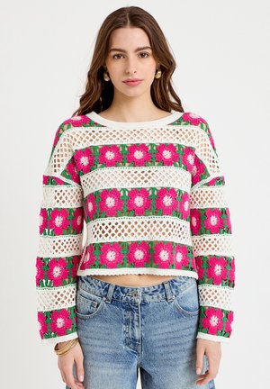 Mujer con una blusa de crochet blanca de manga larga con patrones de flores rosas y verdes, combinada con jeans azules.