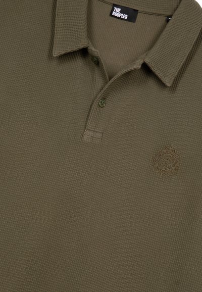 Polo shirt di colore verde oliva con texture, colletto corto, due bottoni ed emblema ricamato sul lato sinistro del petto.