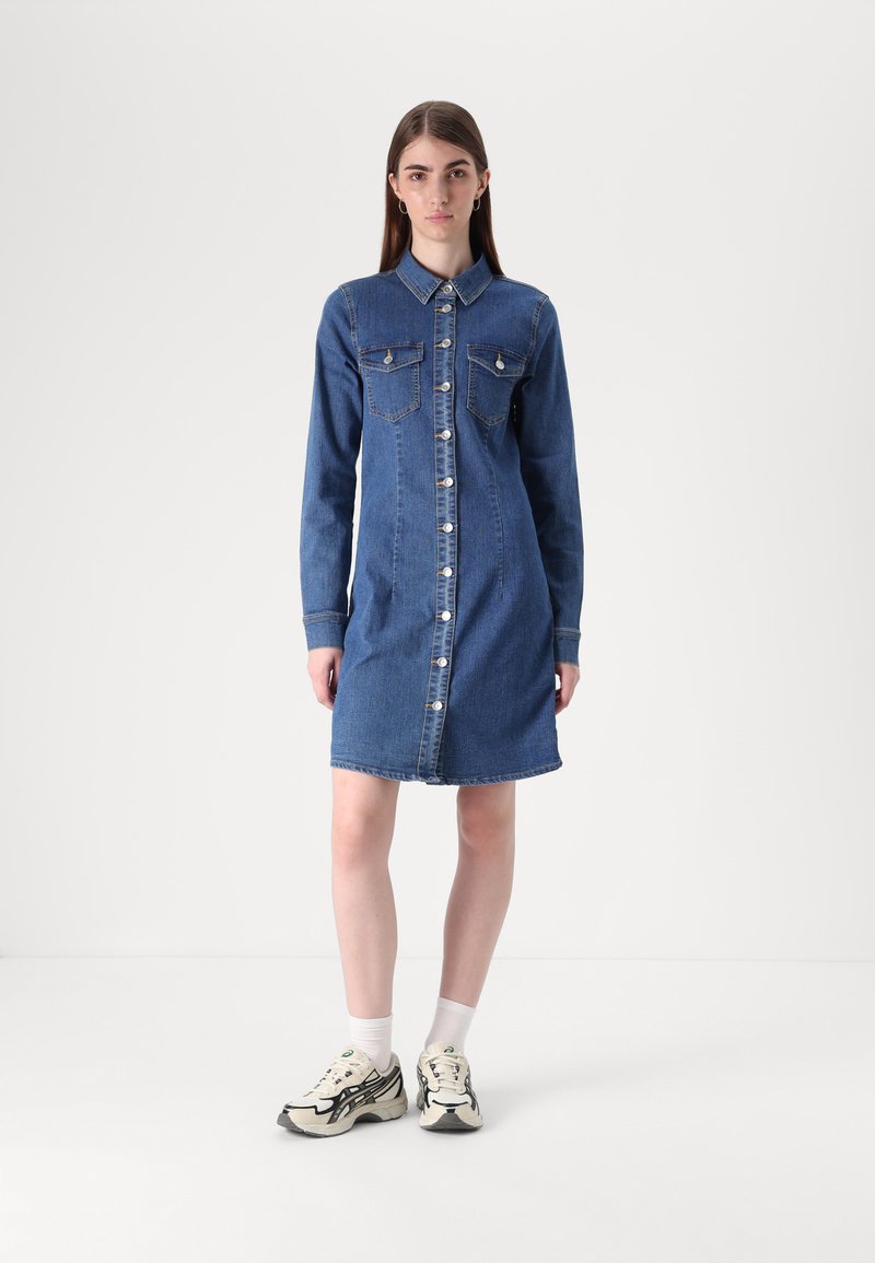 Vestido de camisa de ganga em azul médio com mangas longas, fecho com botões, dois bolsos no peito e corte reto, combinado com sapatilhas de cor clara.