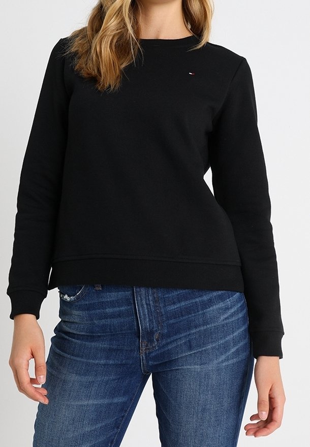 Femme portant un pull noir ajusté à manches longues et col rond, et un jean bleu, avec des cheveux blonds ondulés tombant sur son épaule.