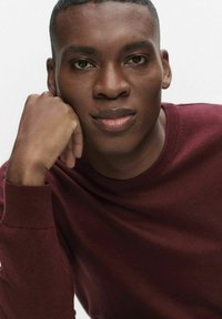 Persoon met kortgeknipt haar, draagt een kastanjebruine trui, rust de kin op de hand met een neutrale gezichtsuitdrukking tegen een lichte achtergrond.