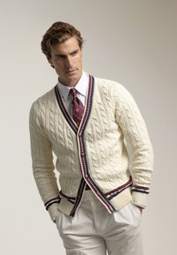 Cárdigan de punto crema con patrón de trenzas, bordes a rayas en navy y burdeos, y botones de madera, llevado sobre una camisa blanca y una corbata.