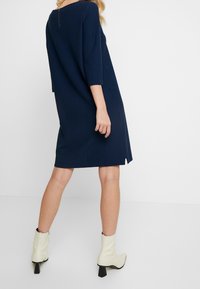 Granatowa, sięgająca kolan sukienka z teksturowanej tkaniny, z rękawami o długości do łokcia, bocznymi rozcięciami oraz suwakiem z tyłu. Połączona z kremowymi botkami za kostkę.