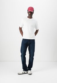 T-shirt bianco a maniche corte, jeans in denim blu scuro e sneakers bianche con accenti neri; modello che indossa un cappellino rosso con visiera curva.