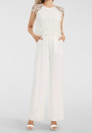 Femme portant un pantalon blanc à jambes larges et un haut sans manches en dentelle blanche à motif floral, debout les mains dans les poches et sandales blanches.