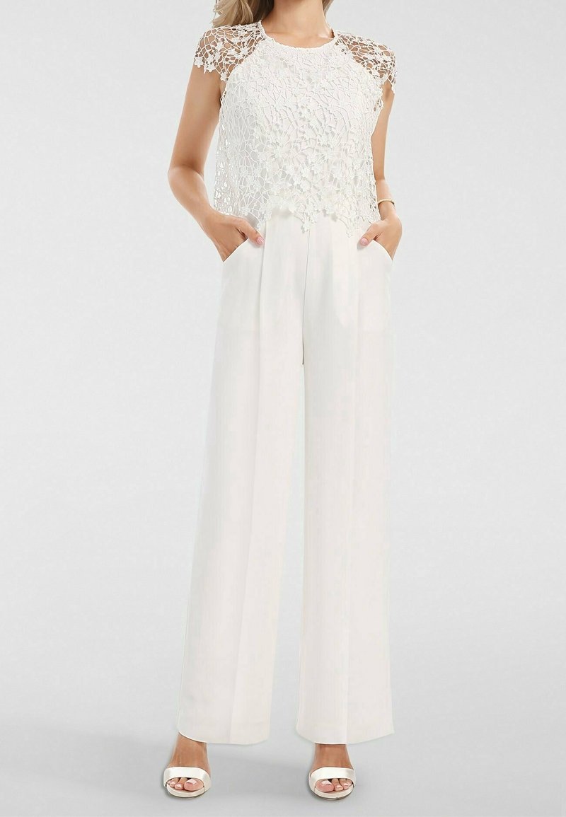 Femme portant un pantalon blanc à jambes larges et un haut sans manches en dentelle blanche à motif floral, debout les mains dans les poches et sandales blanches.