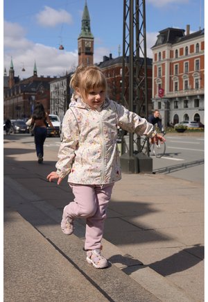 Lyserød mønstret jakke med hætte, lyse lilla bukser og sandaler. Barn går på fortov med bybygninger i baggrunden.