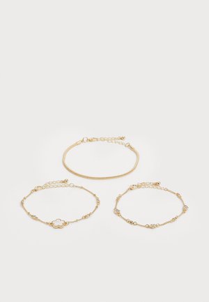 Três pulseiras de ouro: uma com corrente de cobra lisa, uma com pequenos cristais e uma com design floral. Todas possuem fechos ajustáveis.