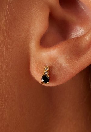 Gouden studs met een druppelvormige zwarte steen onder twee kleine heldere stenen, afgebeeld tegen een close-up van huid.