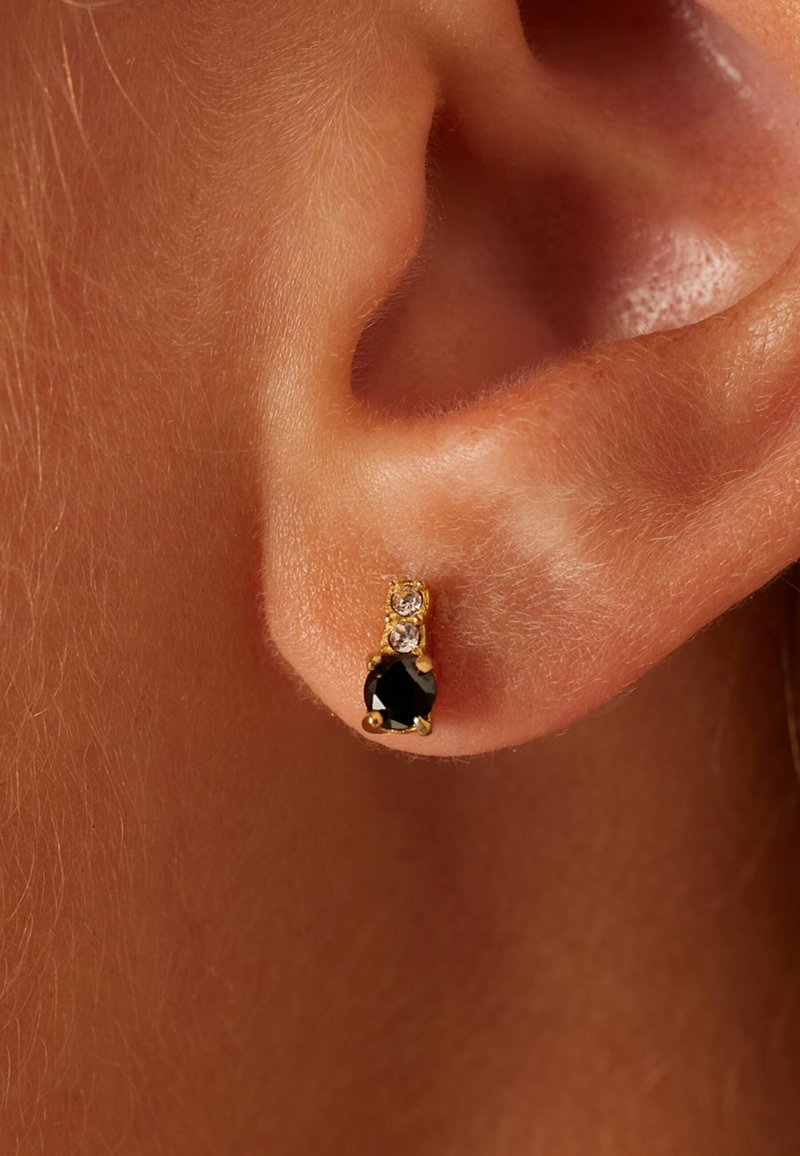 Gouden studs met een druppelvormige zwarte steen onder twee kleine heldere stenen, afgebeeld tegen een close-up van huid.