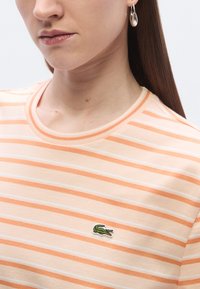 Ριγέ πουκάμισο με κοντά μανίκια από απαλό βαμβάκι σε ροδακινί και λευκό. Διαθέτει λαιμόκοψη και ένα μικρό πράσινο κεντητό λογότυπο στο αριστερό στήθος.