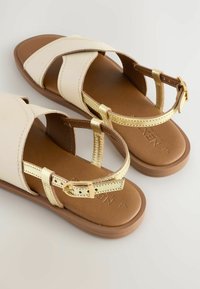Sandali crema con accenti dorati, caratterizzati da cinturini incrociati e chiusure a fibbia, montati su una suola marrone. Realizzati in pelle liscia e materiali sintetici.