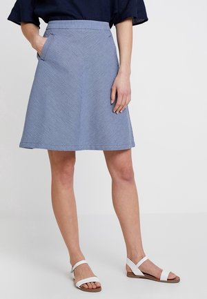 Femme portant une jupe trapèze rayée bleue et blanche avec des poches et des sandales blanches à bout ouvert, debout avec une main dans la poche.