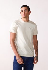 Light beige cotton T-shirt with short sleeves, round neckline, and a small, simple fish graphic on the left chest. Worn with blue jeans.