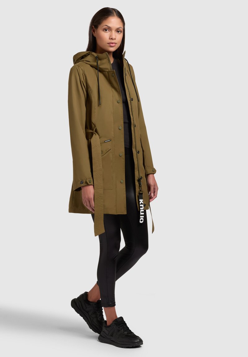 Olivgrüne wasserdichte Jacke mit Kapuze, Reißverschluss, Knöpfen und tailliertem Schnitt. Kombiniert mit schwarzen Leggings und Sneakers. Glatte Textur.