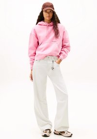 Felpa con cappuccio rosa in cotone con coulisse, jeans bianchi a gamba larga, cappellino marrone e sneakers multicolore con suola testurizzata e dettaglio portachiavi.