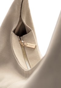 Sac à main en suède beige avec fermeture éclair dorée et étiquette de logo, présentant une texture lisse et une forme arrondie, mis en valeur sous un angle intérieur.