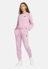 Rosa bomullssweatshirt och matchande sweatpants med elastiskt midjeband, med en svart Nike-logotyp på bröstet och vänster lår, samt vita sneakers.