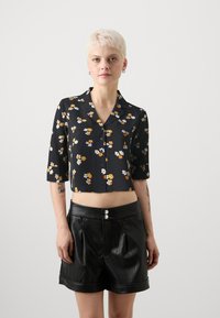 Kuttet svart skjorte med små gule og hvite blomstermønstre, kortermet, og knappelukking; kombinert med høytlivede svarte skinnshorts.
