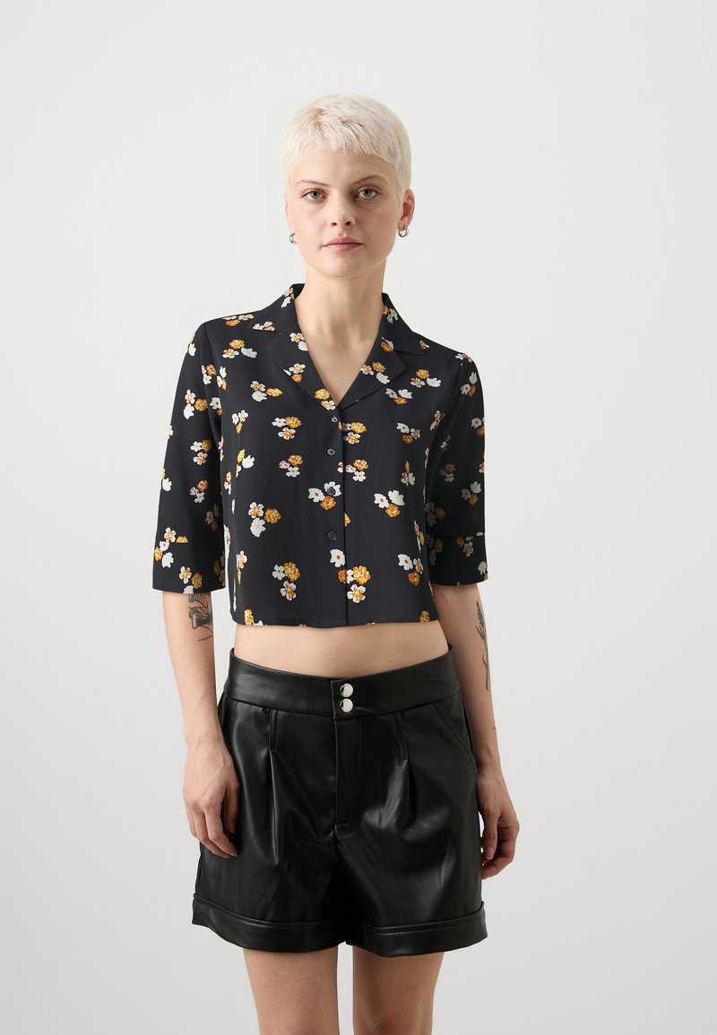 Kuttet svart skjorte med små gule og hvite blomstermønstre, kortermet, og knappelukking; kombinert med høytlivede svarte skinnshorts.