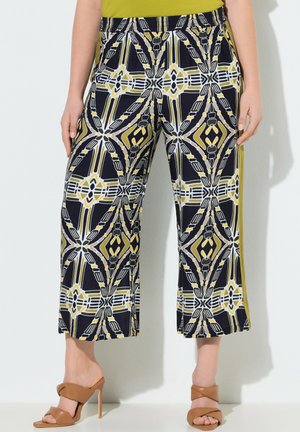 Frau trägt kurze, weit geschnittene Hose mit geometrischem Muster in Schwarz, Weiß und Senfgelb, kombiniert mit beigefarbenen Sandalen mit hohen Absätzen.