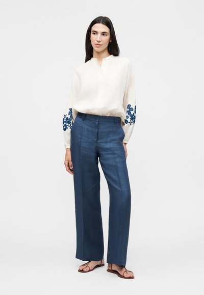 Γυναίκα που φοράει λευκό πουκάμισο με μπλε κεντήματα με λουλούδια στα μανίκια, μπλε παντελόνια με φαρδιά πόδια και ψηλή μέση, και καφέ σανδάλια, στέκεται μπροστά σε απλό φόντο.