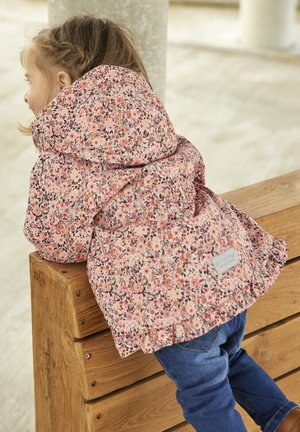 Giacca rosa a fantasia floreale con cappuccio, realizzata in un tessuto morbido e con orlo arricciato, indossata sopra jeans blu. Panchina in legno naturale sullo sfondo.